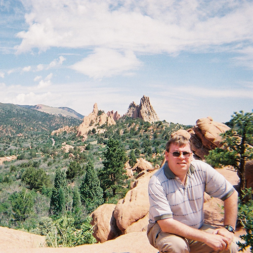 Doug Plester standing on a mountain ledge with mountains and trees in the background in Colorado, USA