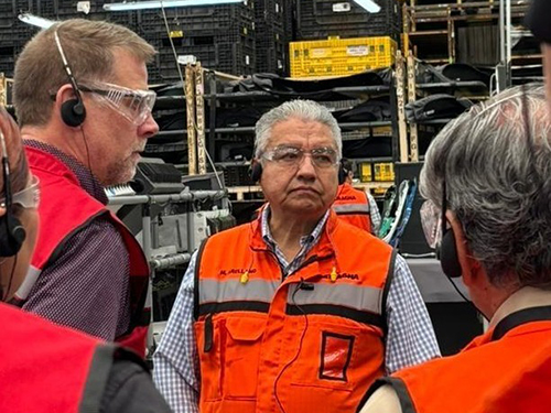 Miguel Arellano and a group of people wearing orange safety vests and protective goggles in a manufacturing facility. They appear focused, possibly in a serious discussion.