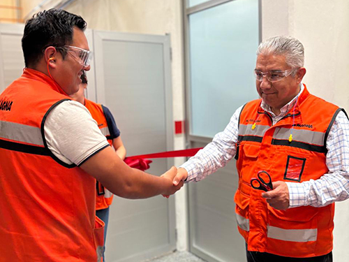 Miguel Arellano and a Magna employee in orange safety vests shake hands in a bright room. One holds scissors, and a red ribbon is partially visible, indicating a ribbon-cutting event.