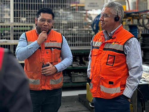 Miguel Arellano and a Magna employee in orange safety vests stand in a manufacturing facility. One listens with a headset, while the other speaks, creating a thoughtful and engaged atmosphere.