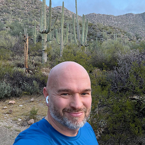 Miroslav Frontz smiling and wearing earbuds outdoors in a natural setting, wearing a blue shirt. The background features cacti, shrubs and rocks.